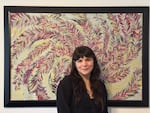 A woman with brown hair smiles for a portrait standing in front of a colorful framed painting that looks like feathers spiraling out from the center of the canvas.