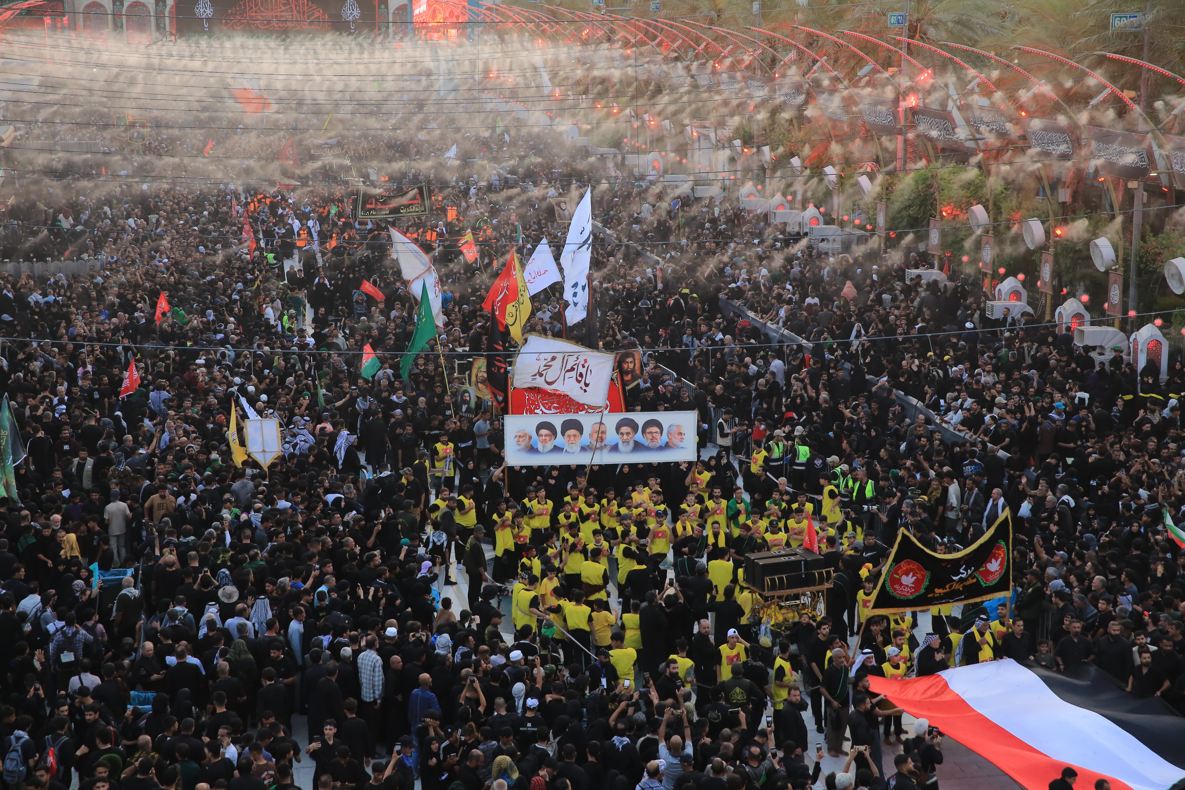 A religious procession moves through dense crowds of pilgrims in the area between the two holy shrines in Karbala, carrying flags, banners and portraits of religious figures during the Arbaeen commemoration.