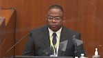 A man in a suit sits in a witness box in a courtroom.
