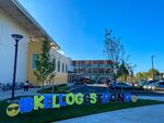 Outside a school building, a handmade sign shows an emoji character wearing sunglasses, a hashtag, and the words "Kellogg strong"