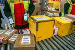 A warehouse worker wearing a fluorescent yellow safety vest uses a scanner to identify cartons of parcels of goods on a conveyor belt in front of transport and delivery lorries in a hangar at DHL s parcel distribution and express freight warehouse on an industrial estate in Valence, France, on 12 December 2024.