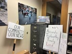 In a room, several picket signs lean up against the wall in the corner, with slogans like "Our Community, Our News," and "Accurate, Trustworthy, Timely, Local." On the top of a filing cabinet sits a photo of people carrying the signs in a parade.