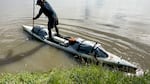 A person stands upright on a long-narrow boat and paddles through murky waters.