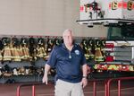 This photo shows Frank Davis, the mayor of Emmitsburg, Maryland, standing in a firehouse. Firefighters' coats and helmets hang in a row on the wall behind him. The front of a firetruck appears on the right side of the frame.
