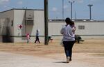 A person carries a small child who rests their head on the adult's shoulder as they walk along a concrete path outside a series of trailers. In the background, other adults and children are seen walking away from a trailer with the medical red cross sign on its side.