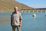 A man with white hair wearing glasses, a plaid light brown and white shirt and a pair of khaki pants stands on the shoreline of a river with a dam behind him.