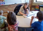 A teacher sits at a table in a classroom in front of three young students as she teaches a reading lesson.