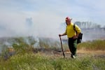 A person wearing a yellow fire shirt, green pants and red fire helmet stands in a smoking field.