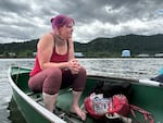 A woman with pink hair sitting in a green rowboat on the river, looking pensively out at the water.