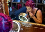 A woman is seen in a mirror with a fancy gold frame, applying red lipstick, with her hair pinned up in rolled curls.