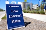 An A-frame sign says "Lennar" and "Low Interest Rates" on it. The letters are white and on a navy blue background. The sign is standing on a strip of ground between a sidewalk and a street curb.