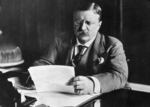 In this black-and-white photo, President Theodore Roosevelt is sitting at his desk circa 1905 and is reading some papers that he's holding.