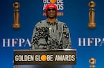 Snoop Dogg wears a red hat and a black and white patterned shirt as he stands at a podium that has the words "Golden Globe Awards written