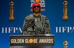 Snoop Dogg wears a red hat and a black and white patterned shirt as he stands at a podium that has the words "Golden Globe Awards written