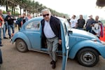 In this 2014 photo, Uruguayan President José Mujica is emerging from the passenger side of his light blue 1987 Volkswagen Beetle. He's wearing gray pants, a light-colored shirt, a dark jacket and sunglasses.