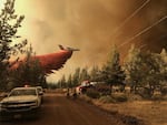 An airplane drops a large plume of reddish pink fire retardant as it flies over trees. A fire truck and police vehicle are in the foreground. Dark heavy wildfire smoke fills much of the sky.