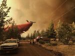 An airplane drops a large plume of reddish pink fire retardant as it flies over trees. A fire truck and police vehicle are in the foreground. Dark heavy wildfire smoke fills much of the sky.