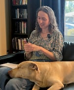 A woman sits on a couch next to a bookshelf, while a one-eyed dog sits calmly beside her and lays its head on her lap.
