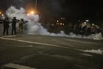 A nighttime image shows law enforcement officers pointing weapons in the direction of protestors who can barely been seen in the shadows, as clouds of tear gas drift in the street.