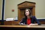 This photo shows Federal Communications Commission Chairwoman Jessica Rosenworcel attending a House subcommittee hearing in 2022. She's sitting at a wooden table and is speaking into a microphone. She's wearing a red shirt and navy blue blazer.