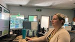 A women sits at a desk with multiple monitors. She wears a headset.