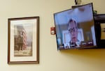 A man in glasses speaks during a video call displayed on a flat screen TV mounted next to a painting of Abraham Lincoln.