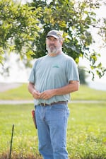 Steve Black stands outdoors. He's wearing blue jeans, a T-shirt and a baseball cap. Green grass is in the background, and tree branches with green leaves hang in the background.