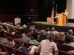 U.S. Rep. Val Hoyle answers questions at an Aug. 20, 2025 town hall in Eugene while a protestor holds a banner.