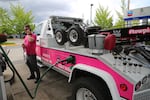 A tow truck driver stands outside next to their truck and talks on a cell phone while the truck is filled with gasoline.