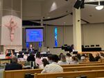 A pastor speaks before a congregation in a church while standing in front of a crucifix.