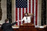An aide hands Nancy Pelosi something while the speaker stands at the lectern in the House Chambers. An American flag hangs vertically behind her.
