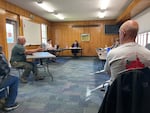 A man sitting on the left side of the room speaks at a table facing Union County commissioners sitting on the right side. In the foreground, a man watches the proceedings with a shirt that states "Move Oregon's Borders" on the back.