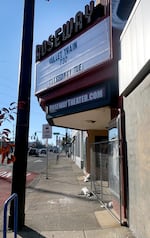 A movie theater marquee juts above a sidewalk, while a chain link fence blocks the entryway and ticket window.
