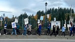 A group of people holding signs picket on an overpass above a highway on a cloudy day.