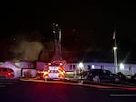 A fire engine with its ladder up is seen parked outside a residential complex with a dark sky. The roof of the building has been burned and damaged, as smoke emits from it.