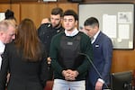 Luigi Mangione appears in court, wearing a bullet proof vest and green sweater over a while collared shirt with his hands clasped in handcuffs.