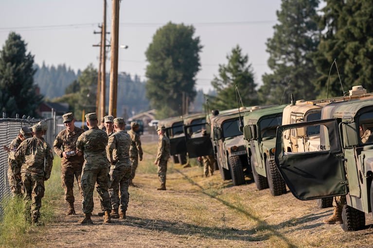 National Guard members in uniforms stand together and talk next to a row of parked military vehicles.