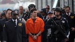 Luigi Mangione, wearing a bright orange jumpsuit, is surrounded by NYPD officers, including some with guns.