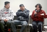 A young woman talks through microphone her other hand gesturing for effect as she speaks. Seated next to her are two men, with the one closest to her folding his arms.