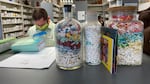 A masked pharmacist is looking down, standing behind a counter that has three decorative jars full of pills that are layered by color.