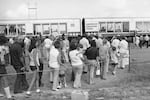This black-and-white photo shows two cars of the American Freedom Train on June 20, 1975, in Archbold, Ohio. The cars have big windows, and inside are historical artifacts, such as an early automobile. In the foreground, people wearing contemporary clothing, such as bell-bottom pants, are lined up.