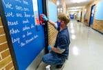 a man wearing a mask, squatting in front of a bright blue classroom door, using a red rag to wipe the door and doorknob down