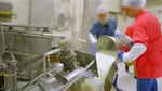 Two workers in hair nets and scrubs pour a bucket of sour cream into a large drum at the start of a batch of beef stroganoff for freeze drying.