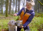 A white woman with blonde hair and blue eyes smiles as she pulls a rehydrated cheesy fork full of food from a freeze-dried meal pouch while cooking in a forest. Her golden retriever stands next to her, looking eager for a bite.