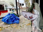 A person wrapped in a sleeping bag holds a plastic poncho and umbrella while standing next to a tent on a city sidewalk outside.