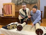 A man reads from the Torah using a traditional pointer to follow the words, while the rabbi looks on, holding a microphone for the reader.