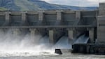 Water gushes through a concrete dam, with hills visible in the distant background.