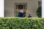 In this photo, President Trump is waving with one hand while outside the White House. A row of hedges is in the foreground.