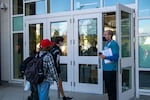 Students approach a door while an adult stands outside wearing a mask and holding a radio and a clipboard.