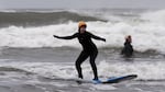 A woman wearing a wetsuit stands upright on a surfboard while riding a small wave.